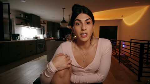 Video of a woman with dark hair in a bun, wearing a pink top, sitting in a modern kitchen with a bar and warm lighting.