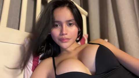 Video of a Latina woman with medium skin tone, long black hair, and brown eyes, wearing a black bra, lying on a bed with beige and red striped pillows and beige curtains in the background.
