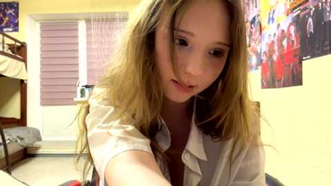 Video of a young Caucasian woman with long, blonde hair, wearing a white shirt, sitting on a bed in a cozy bedroom with posters and a window with purple blinds.
