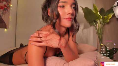 Media: Video of a slender, light-skinned woman with dark hair, lying on her stomach on a bed. She wears black lingerie, revealing her small breasts. The room has a beige wall, a green plant, and a nightstand with a bottle.