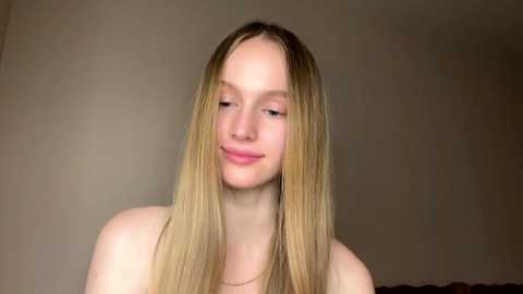 Video of a young, fair-skinned woman with long, straight blonde hair, wearing a pink top, smiling slightly, against a plain beige wall background.