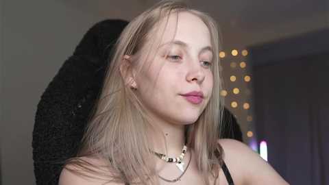 Media: Video of a young, fair-skinned woman with long, straight blonde hair and a serene expression, wearing a black top and a gold necklace, against a softly lit, blurred indoor background.