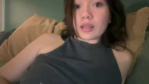 Video of an Asian woman with shoulder-length dark hair, fair skin, and wearing a black sleeveless top, reclining on a bed with beige pillows.