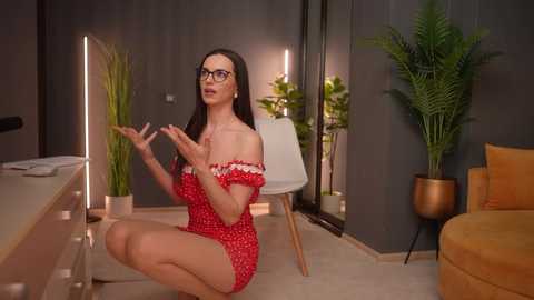 Media: Video of a woman with long brown hair, wearing glasses and a red off-shoulder dress, sitting cross-legged on a beige carpet in a modern, dimly-lit room with green plants, a white chair, and a yellow couch.