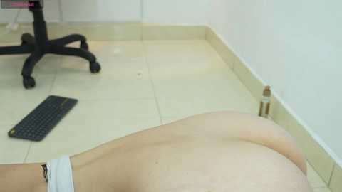 A video of a naked person lying on a tiled floor in an office, with a black keyboard nearby. The person's buttocks are prominent, and their skin is light.
