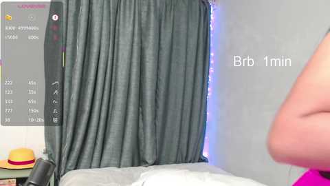 Video of a bedroom with a grey curtain, a white bed, and a pink hat on a nightstand, displaying \"Bro 1 min\" on a screen.