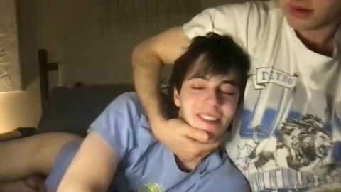 A video shows a young man with fair skin and dark hair in a blue T-shirt being held by another man in a white graphic tee, with a wooden cross and lamp in the background.