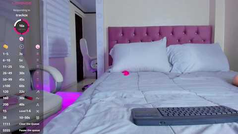 Media: A video of a modern bedroom with a pink tufted headboard, white bedding, and a black keyboard on the bed. The room is dimly lit with purple ambient lighting.
