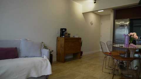 A cozy, minimalist bedroom with a wooden dresser, a white bed, and a small table with a vase of flowers; light beige walls and a polished concrete floor.