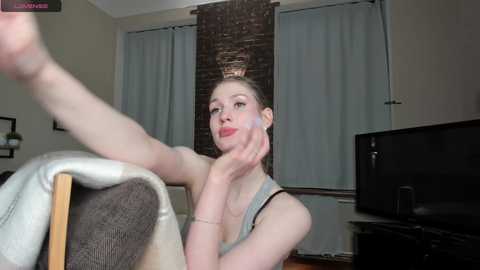 Media: Video of a fair-skinned woman with blonde hair in a bun, wearing a grey tank top, sitting on a beige couch, lifting her leg. Background includes a brick wall, white curtains, and a TV.