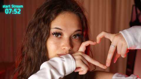 Video of a young woman with long brown hair, brown eyes, and fair skin, wearing a white top, making a heart shape with her hands, against a blurred background.