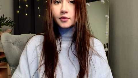 Video of a young woman with long, straight brown hair, wearing a light blue turtleneck sweater, indoors. Background includes a green plant, a gray couch, and string lights.