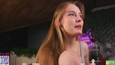 Video of a young woman with long, straight blonde hair, wearing a grey tank top, looking upward, in a dimly lit room with greenery and a QR code watermark.