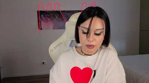 A video of a young woman with short black hair, wearing a white sweatshirt with a red heart, seated in a beige gaming chair. The background features a wall with pink neon lights and a large eye painting.