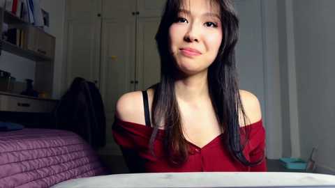 Video of an Asian woman with long black hair, wearing a red off-shoulder top, sitting in a dimly lit bedroom with a purple quilted bed and a cluttered desk in the background.