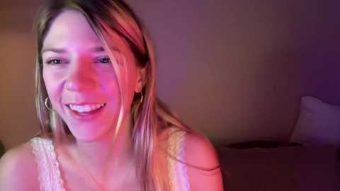 Video of a smiling, light-skinned woman with long, straight blonde hair, wearing a white lace tank top and large hoop earrings, in a dimly lit room with purple and pink lighting.