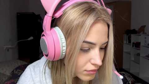 Video of a young woman with long blonde hair, wearing pink cat ear headphones and a white robe, in a dimly lit bedroom.