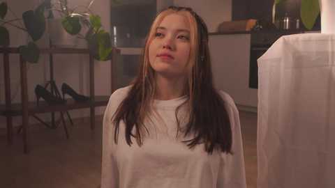 Video of a young Asian woman with long, dark hair in a white shirt, standing indoors, looking upward. Background includes potted plants, kitchen items, and wooden floors.