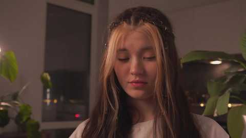 Video of a young girl with long, dark hair and blonde streaks, wearing a white top, standing in a dimly lit room with potted plants and a window in the background.