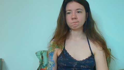 Media: Video of a young Caucasian woman with long brown hair, wearing a black lacy bralette and a colorful patterned robe, standing against a plain light blue wall.