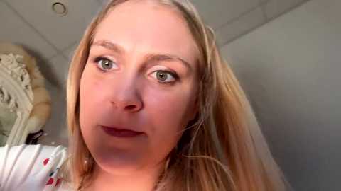 Video of a fair-skinned, light-haired woman with green eyes, wearing a white blouse with red polka dots, smiling slightly. Background features an ornate mirror and neutral-colored walls.