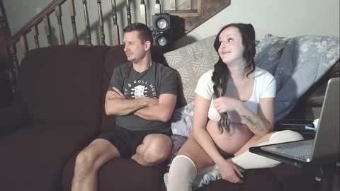Video of a couple sitting on a dark brown couch in a cozy living room, watching TV. The man, muscular, wears a dark T-shirt and shorts, while the woman, slender, wears a white top and white knee-high socks, holding a laptop.