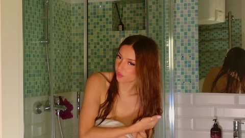 Video of a slim, fair-skinned woman with long, wet, brown hair, wearing a white towel, standing in a modern, green-tiled bathroom with a glass shower and mirror.