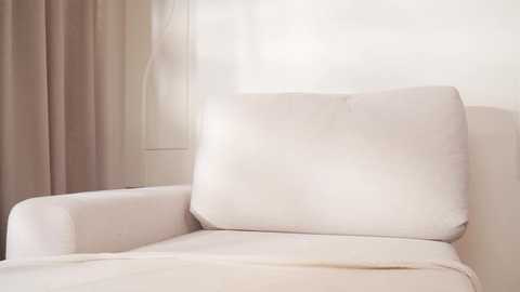 A video of a beige armchair with a plush, soft cushion against a plain white wall and a sheer, light-colored curtain. The image is well-lit with natural light, emphasizing the smooth texture and cozy feel of the furniture.