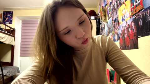 Media: Video of a young Asian woman with long brown hair, wearing a beige long-sleeve top, leaning over a bed in a colorful, cluttered bedroom.