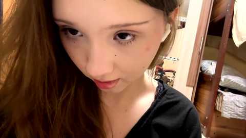 Media: A video of a young, fair-skinned woman with long, straight brown hair, wearing a black top, and looking directly at the camera, taken indoors in a bedroom with visible bed and furniture.