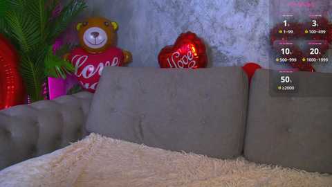 A video of a plush grey couch with a red throw blanket and a plush teddy bear with \"I Love You\" written on it. The background has a textured, silver wall with a red heart-shaped balloon.