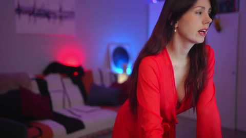 Media: Video of a woman with long brown hair in a red dress, leaning forward in a dimly lit bedroom with a bed and red lights.