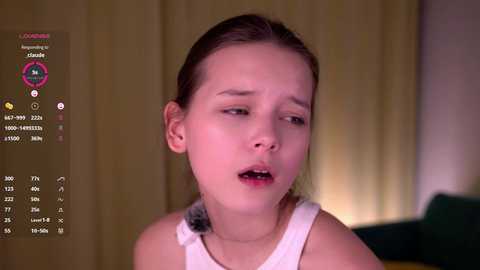 Video of a young Caucasian girl with fair skin and brown hair tied back, wearing a white sleeveless top. She appears distressed, eyes half-closed, mouth open. Background features beige curtains and green furniture.