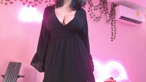 Video of a curvy woman with light skin and shoulder-length, wavy black hair, wearing a black, V-neck dress, standing in a pink-themed room with a chair, plants, and heart-shaped lights.