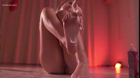 A video of a nude woman with fair skin, reclining on a tiled floor, her feet covered in white foam, red lighting creating a sensual ambiance.
