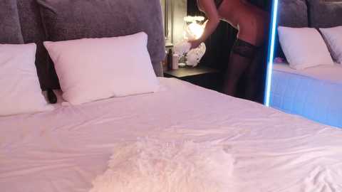 Video of a woman in black lingerie adjusting her bra on a bed with white sheets and a fluffy pillow.