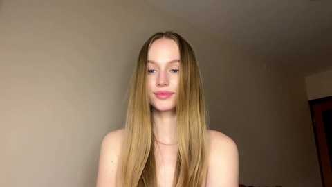Video of a young, slender, light-skinned woman with long, straight, blonde hair parted in the middle, standing against a plain beige wall, wearing no visible clothing.