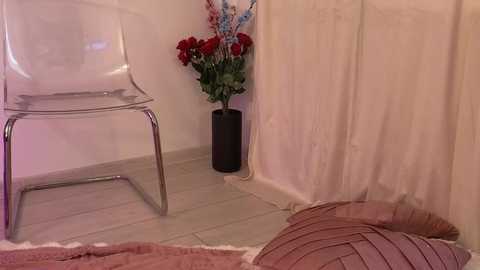 Video of a modern, minimalist room with a clear chair, a vase of fake flowers, and a pink knitted cushion on a beige tiled floor.