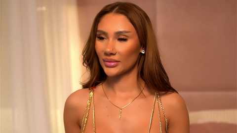 Video of a woman with long brown hair, wearing a gold chain necklace, pink lipstick, and a sleeveless gold top, set against a blurred, warm-toned background.