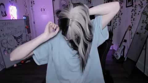 Video of a person with long, platinum-blonde hair and black roots, wearing a light blue t-shirt, adjusting their hair in a bedroom with purple lighting, white walls with anime-themed posters, and a guitar.