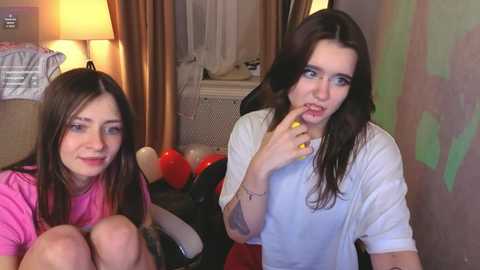 Video of two young women with long hair, one in a pink top, the other in white, seated on a couch, surrounded by balloons and a lamp in a cozy room.