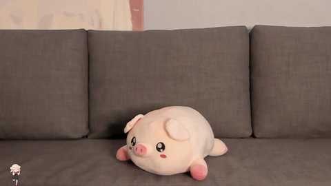 Video of a plush piglet toy with pink fur, white ears, and a small pink flower on its head, lying on a gray fabric sofa against a beige wall.