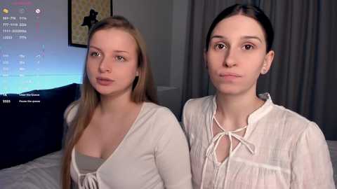 Video of two young women with fair skin and straight, blonde hair, dressed in casual, light-colored tops, standing indoors.