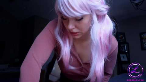 Video of a young woman with light skin, pink hair, and a pink shirt leaning forward in a dimly lit room with a dark background.