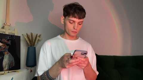 Video of a young man with short dark hair, wearing a white t-shirt, focused on his smartphone, in a dimly lit room with a rainbow reflection.