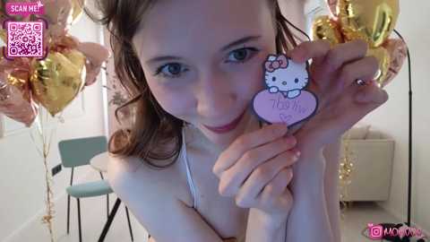 Video of a young woman with light skin, long brown hair, and blue eyes, holding a Hello Kitty pin with \"Thanks\" written on it. She's in a room with gold balloons and a teal chair in the background.