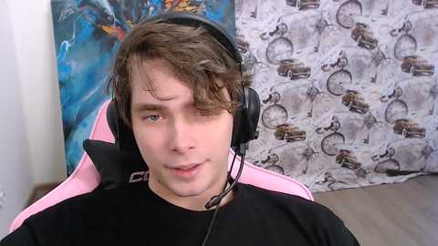 A video of a young, light-skinned man with curly brown hair wearing black headphones and a black shirt, seated in a pink gaming chair. The background features abstract blue and white art on the left and a white curtain with brown bicycle pattern on the right.