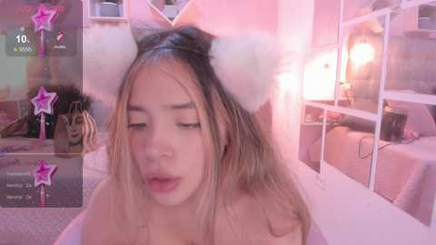 A video of an Asian woman with long, blonde hair, wearing a white cat ear headband, and pink lipstick, in a cozy bedroom with pink lighting, looking seductive.