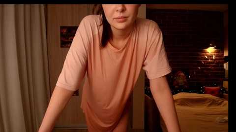 Media: Video of a young woman with light skin, brown hair, and glasses, wearing a loose peach T-shirt, leaning forward in a cozy bedroom with brick wall, bed, and soft lighting.