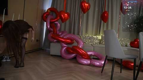 Video of a room decorated with large, metallic heart-shaped balloons in red and pink, suspended from the ceiling. A white chair and a person in black attire are visible.